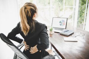 A young business woman seated at her office desk clutches at her back for "Should I Visit Urgent Care for Back Pain" blog.