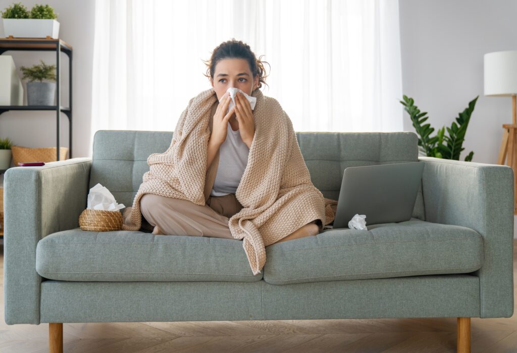 A young woman sits on her couch, draped in a blanket, blowing her nose, with a box of tissues at the ready for "Should I Go to Urgent Care for a Cold" blog.