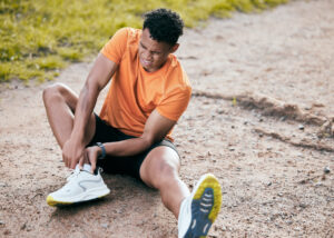 A man in an orange shirt and black shorts sits on a sidewalk gripping his ankle for the should-I-go-to-urgent-care-for-a-sports-injury blog.
