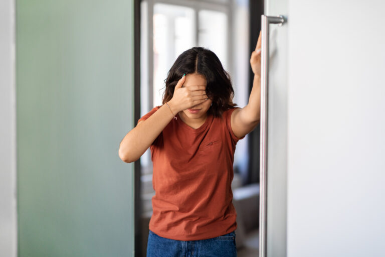 A young woman in a dark orange shirt suffers from dizziness and headache, bracing herself against the wall and holding her head for urgent-care-for-carbon-monoxide-exposure blog.