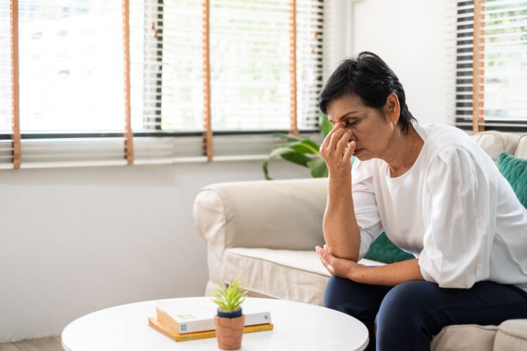 A senior Asian woman sits alone on the couch, holding the bridge of her nose due to headache for urgent-care-for-sudden-high-blood-sugar blog.