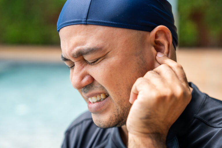 A man wearing a swim cap pinches his ear with a grimace, suggesting ear pain or discomfort after swimming for urgent-care-for-swimmers-ear blog.