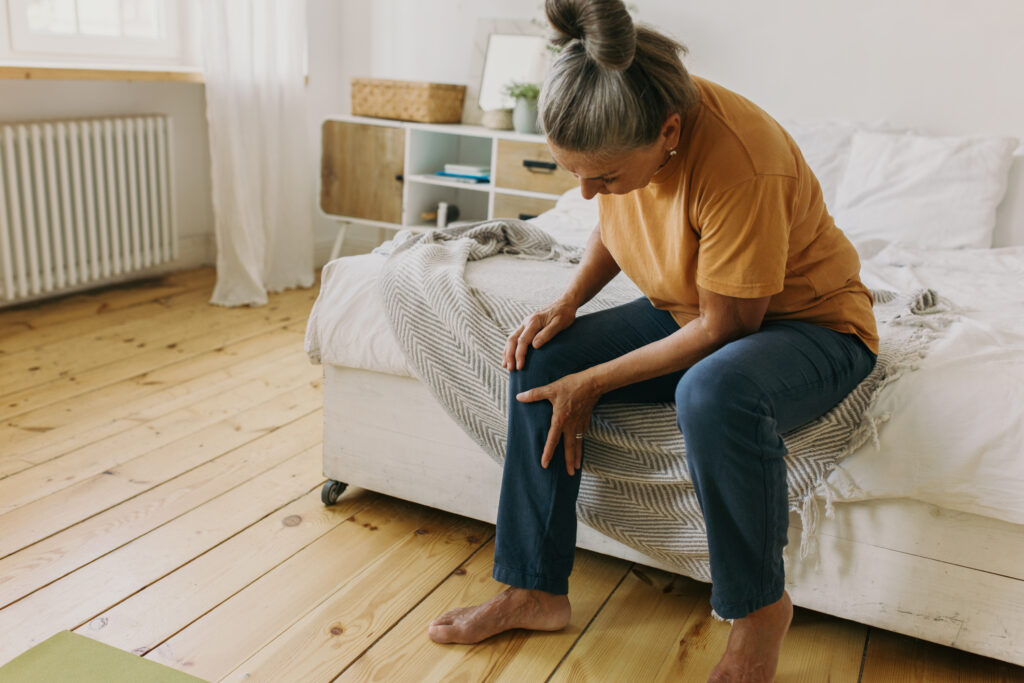 A senior woman in an orange shirt sits on the side of her bed, massaging her leg for urgent-care-for-severe-muscle-cramp blog.